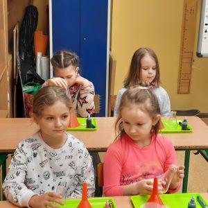 Na zdjęciu widać cztery uczennice klasy pierwszej oraz drugiej, dziewczynki siedzą przy stołach na których przygotowane są na tacy akcesoria do wykonywania doświadczeń.