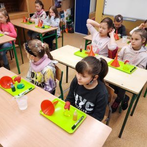 Na fotografii widać uczniów klasy pierwszej oraz drugiej siedzących przy ławkach. Przed nimi leżą tace z akcesoriami do przeprowadzenia doświadczeń między innymi lejek, szklana kolba, barwnik.