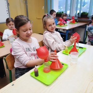 Na zdjęciu uczennice klasy drugiej wypełniają balon ciepłym powietrzem. Naciągnięty na szklane naczynie balon pod wpływem temperatury wypełnia się powietrzem.