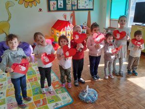Fotografia przedstawia grupę przedszkolaków trzymających serduszka z odciskami swoich dłoni. Przed dziećmi leży torba z nakrętkami zebranymi w akcji ekologicznej.
