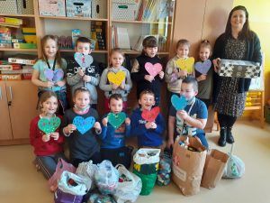 Na fotografii grupa uczniów klasy pierwszej wraz z wychowawczynią prezentują kolorowe serduszka przygotowane dla chorej Ani. Przed dziećmi stoją torby z zebranymi nakrętkami. Wychowawczyni trzyma kartoniki z pączkami.