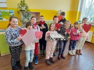 Fotografia przedstawia dziecięciu uczniów klasy drugiej. Dzieci trzymają w rękach czerwone serduszka. Jedna z dziewczynek ma kartonik z pączkami.