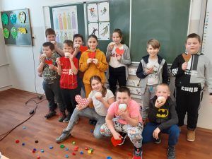 Na fotografii dziewczynki i chłopcy z klasy trzeciej zajadają pączki. Kilkoro trzyma w rękach czerwone serduszka. Dzieci znajdują się w sali lekcyjnej.