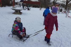 Fotografia przedstawia dwie dziewczynki bawiące się w na ośnieżonym placu zabaw. Jedna siedzi na sankach, druga ją ciągnie. W tle widać inne bawiące się dzieci.