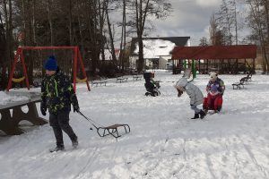 Na fotografii grupa uczniów klasy drugiej bawi się na zaśnieżonym placu zabaw. Chłopiec ciągnie sanki. Za nim dziewczynki ciągną się na sankach.