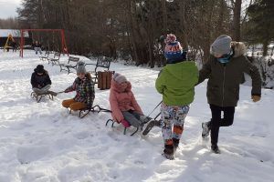 Na zdjęciu uczniowie klasy trzeciej na zaśnieżonym placu zabaw bawią się w kulig. Dwaj chłopcy ciągną sanki, na których siedzi troje dzieci.