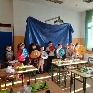 Na fotografii stoją uczniowie na tle niebieskiej płachty każdy z nich trzyma w rękach planetę w takiej kolejności w jakiej występują one w Układzie Słonecznym.