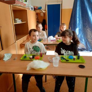 Na zdjęciu widać uczniów klasy pierwszej, na ich ławkach widać tace z barwnikami, watą, pipetami, słoikami i innymi przyborami potrzebnymi na warsztatach. 