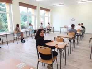 Zdjęcie przedstawia uczniów w sali egzaminacyjnej. Jeden z nich odwrócony i uśmiechnięty.