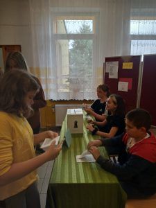 Zdjęcie przedstawia komisję wyborczą i uczennicę wypełniającą kartę.