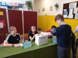 Zdjęcie przedstawia ucznia wrzucającego swoją kartę do głosowania do urny wyborczej.