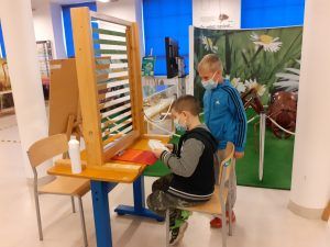 Dwaj chłopcy rozwiązują zadania. Jeden siedzi przy stoliku na którym znajduje się przyrząd iluzji optycznej. Drugi chłopiec stoi i przygląda się pracy kolegi.