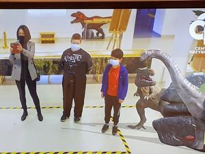 Dziewczynka i chłopiec oraz nauczycielka robiąca zdjęcie. Po prawej stronie znajdują się dwa dinozaury.