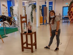 Stojąca dziewczyna a obok niej  na krześle siedzi miniaturka drugiej dziewczynki. Fotografia powstała w wyniku zastosowania iluzji optycznej.