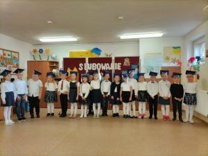 W półkolu stoją wszyscy uczniowie klasy pierwszej z biretami na głowach.