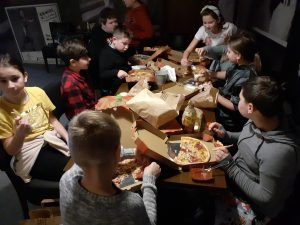 Na fotografii 8 grupa dzieci siedzi przy dużym stole, na którym rozłożone są kartoniki z pizzą, leżą sosy, butelki z napojami. Dzieci jedzą posiłek.