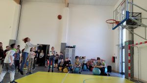 grupa uczniów dopingujących nauczycielce rzucającej piłkę do kosza