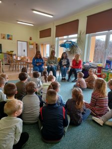 Na zdjęciu przedszkolaki siedzą na dywanie i słuchają bajek czytanych przez starsze koleżanki.