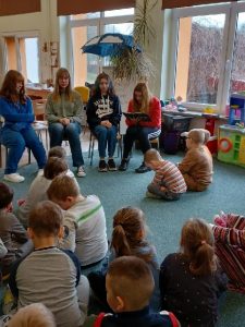 Na zdjęciu przedszkolaki siedzą na dywanie i słuchają bajek czytanych przez starsze koleżanki.