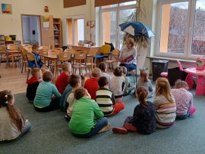 Na zdjęciu przedszkolaki siedzą na dywanie i słuchają bajek czytanych przez Panią Ewę Mulawę