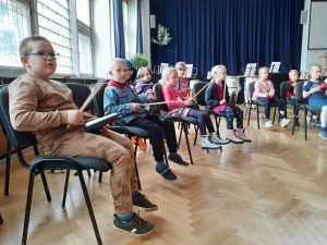 Dzieci siedzą na krzesełkach i podczas zajęć muzycznych grają na małych instrumentach perkusyjnych.