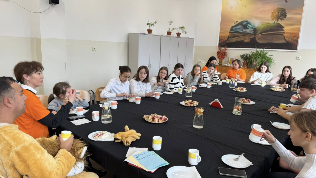 Grupa dzieci i dorosłych siedzi przy dużym stole nakrytym czarnym obrusem, jedząc poczęstunek i rozmawiając. 