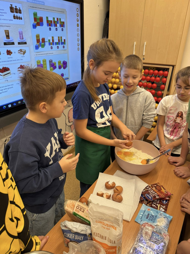 Grupa dzieci w klasie wspólnie przygotowuje ciasto na pierniki, mieszając składniki w dużej misce na stoliku pełnym produktów do pieczenia.