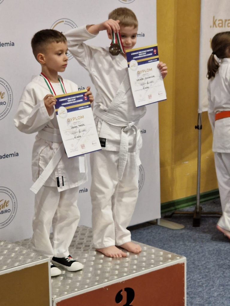Dwóch chłopców w strojach karate stoi na podium, trzymając w rękach dyplomy. Na szyjach mają zawieszone brązowe medale za zajęcie trzeciego miejsca w zawodach.
