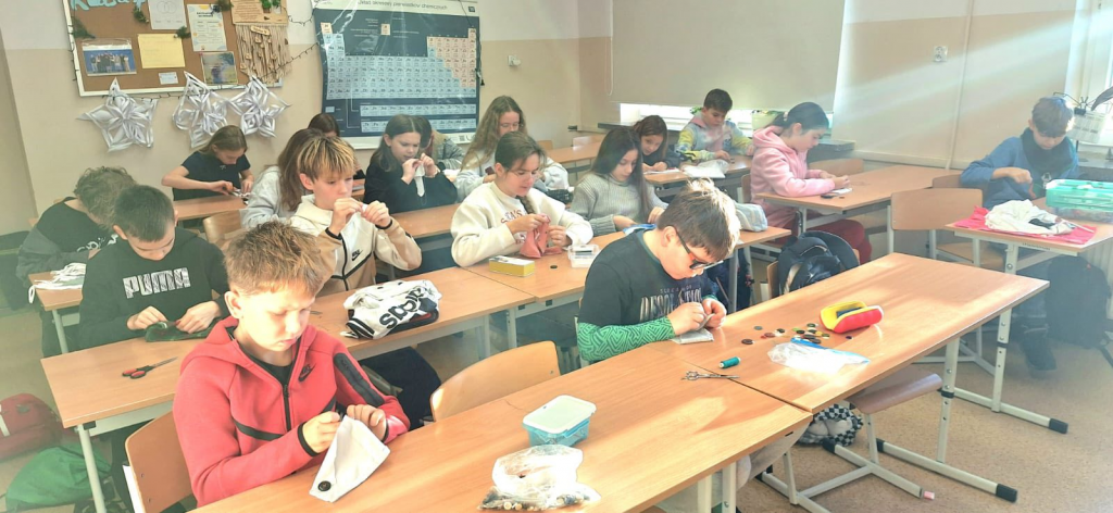 Uczniowie siedzą w szkolnej klasie przy ławkach i skupieni wykonują prace ręczne, szyjąc lub przypinając guziki z użyciem igieł, nici i materiałów.