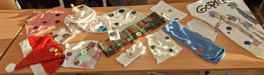 Na szkolnej ławce leżą kolorowe kawałki materiałów i ręcznie przyszyte guziki – efekty pracy uczniów podczas zajęć z szycia.