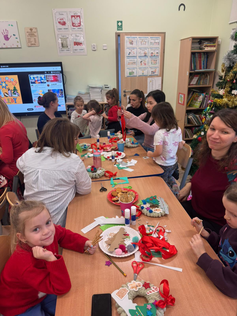 Grupa dzieci i dorosłych siedzi przy połączonych stołach w sali przedszkolnej i wspólnie wykonuje prace plastyczne. Na stołach leżą nożyczki, klej, kolorowy papier, wstążki, pompony i gotowe ozdoby, a także talerzyki z ciasteczkami. W tle widać tablicę multimedialną, regał z książkami oraz udekorowaną choinkę. Dzieci skupiają się na pracy, a jedna dziewczynka z przodu uśmiecha się do aparatu.