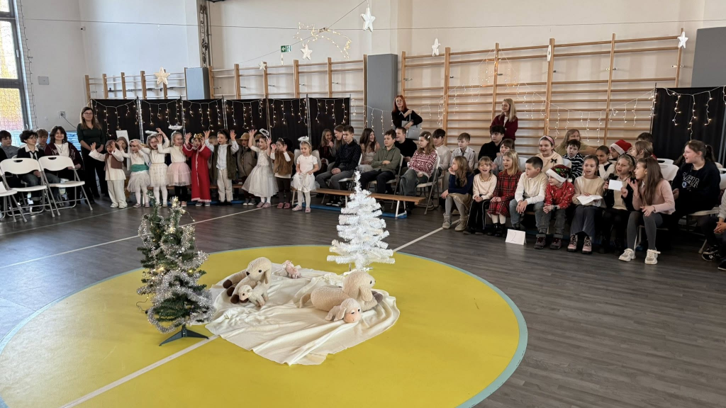 Grupa dzieci w świątecznych strojach stoi i siedzi w sali gimnastycznej podczas szkolnego występu. Na środku sali znajduje się dekoracja z małymi choinkami, jasnym materiałem i pluszowymi owieczkami, nawiązująca do sceny bożonarodzeniowej. W tle widać publiczność złożoną z uczniów i nauczycieli oraz świąteczne dekoracje i światełka.