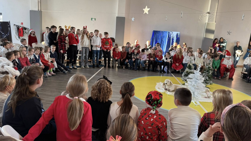 Grupa dzieci w świątecznych strojach stoi i siedzi w sali gimnastycznej podczas szkolnego występu. Na środku sali znajduje się dekoracja z małymi choinkami, jasnym materiałem i pluszowymi owieczkami, nawiązująca do sceny bożonarodzeniowej. W tle widać publiczność złożoną z uczniów i nauczycieli oraz świąteczne dekoracje i światełka.