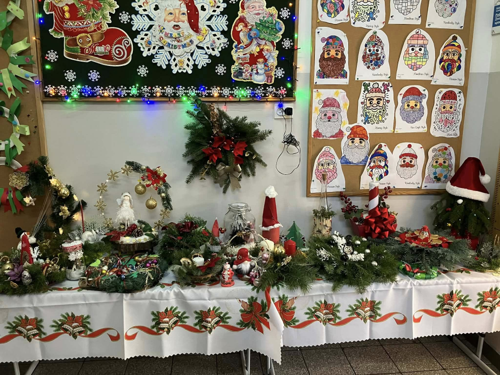 Świątecznie udekorowany stół w sali szkolnej lub przedszkolnej, przykryty białym obrusem z bożonarodzeniowym wzorem. Na stole znajdują się liczne stroiki z gałązek świerku, świeczniki, figurki Mikołajów, ozdoby świąteczne, bombki, wstążki i lampiony. W tle widać tablice dekoracyjne z motywami świątecznymi, kolorowe lampki oraz prace plastyczne dzieci przedstawiające Mikołajów. Całość tworzy ciepłą, radosną atmosferę świąt Bożego Narodzenia.