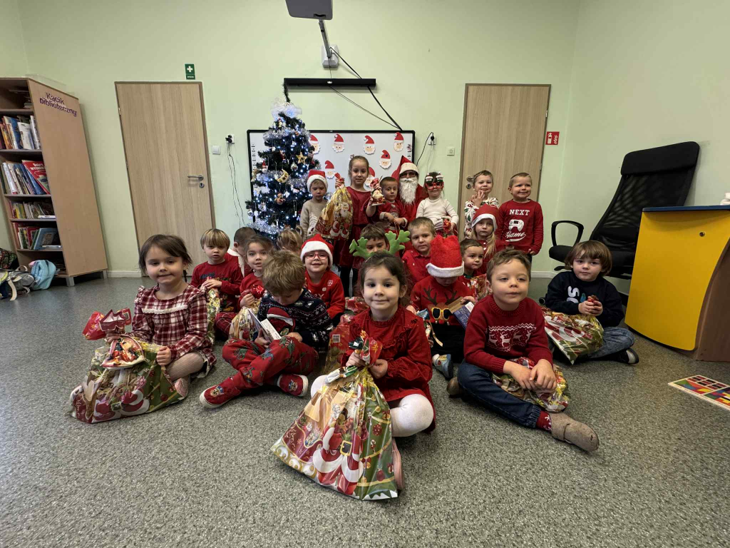 Grupa małych dzieci siedzi z Mikołajem na podłodze w klasie, każde z nich trzyma świąteczną paczkę. Dzieci ubrane są w czerwone i świąteczne stroje. W tle stoi ozdobiona choinka oraz tablica z rysunkami Mikołaja. Wokół widać elementy sali przedszkolnej, takie jak regał z książkami i biurko nauczyciela. Wszystkie dzieci patrzą w stronę aparatu.