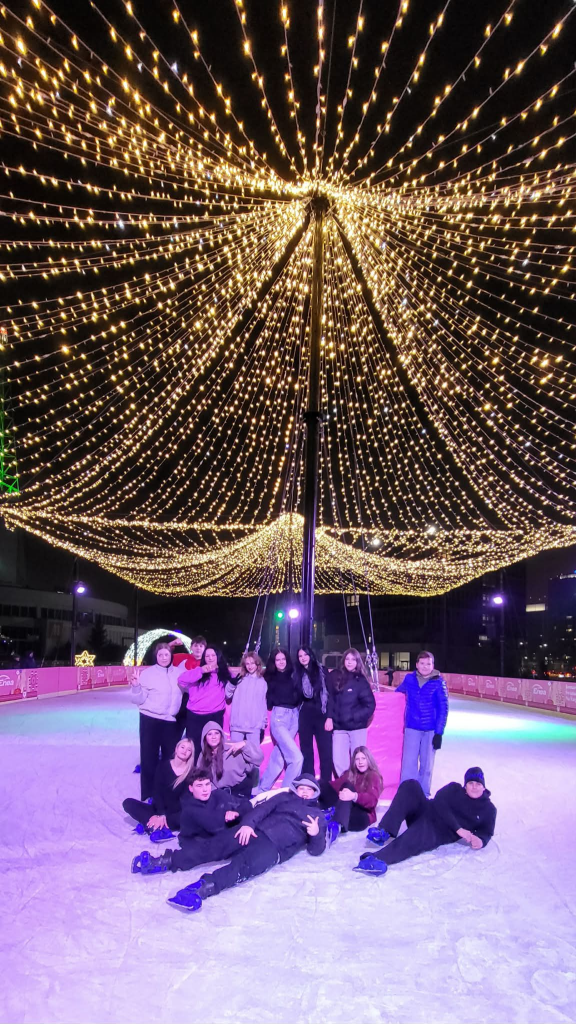 Grupa uczniów pozuje na lodowisku pod świątecznymi świetlnymi dekoracjami.