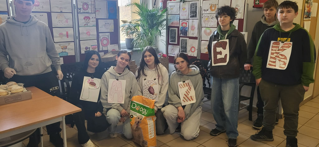 Dzieci pozują do zdjęcia z karmą dla zwierząt. Mają na sobie kartki z literami, które układają się w wyraz  ,,pączek". W tle widać plakaty z pączkami.