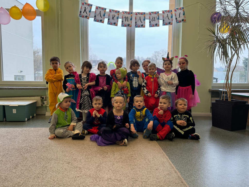 Dzieci w przebraniach karnawałowych pozują do zdjęcia. W tle są balony i girlanda z napisem ,,karnawał".