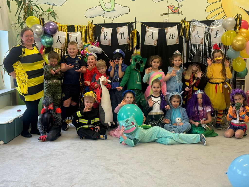 Dzieci w przebraniach karnawałowych pozują do zdjęcia. W tle są balony i girlanda z napisem ,,karnawał".