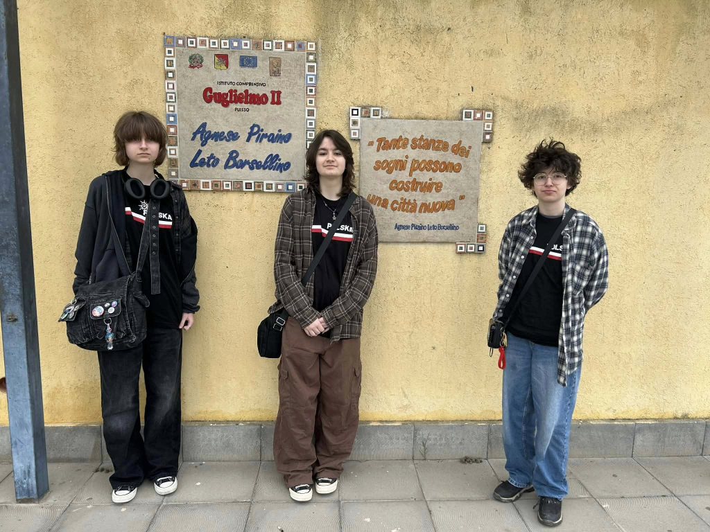 Trzy osoby stoją przed żółtą ścianą budynku szkolnego. Za nimi znajdują się dwie tablice z nazwą szkoły oraz cytatem po włosku.