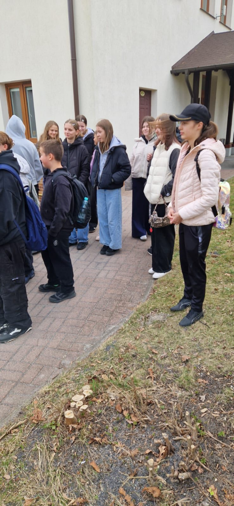 Grupa uczniów stoi przed budynkiem i słucha, przygotowując się do wyjścia.