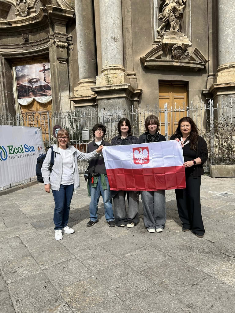 Trzy uczennice trzymają polską flagę z białym orłem, a po bokach stoją dwie nauczycielki. Wszyscy pozują do zdjęcia przed zabytkowym kościołem z kolumnami i rzeźbami.