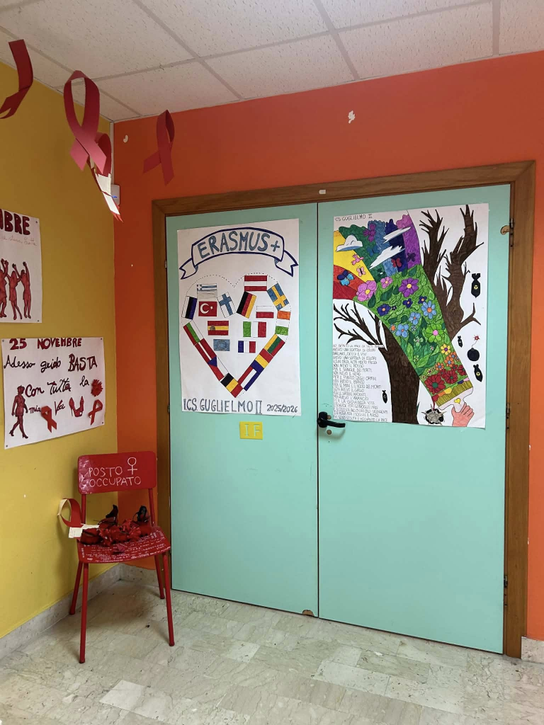 Korytarz szkolny z jasnoniebieskimi drzwiami klasy. Na drzwiach wiszą dwa duże plakaty uczniowskie: jeden z napisem „Erasmus+” i sercem złożonym z flag różnych krajów, drugi przedstawiający kolorowy krajobraz z drzewem i kwiatami. Po lewej stronie stoi czerwone krzesło z napisem „Posto occupato” oraz czerwonymi wstążkami – symbol akcji przeciw przemocy wobec kobiet. Na ścianach znajdują się również czerwone wstążki i plakaty związane z dniem 25 listopada.