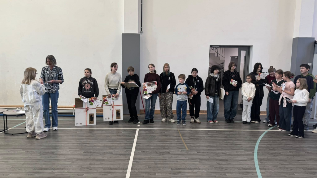 Grupa uczniów stoi w sali gimnastycznej podczas apelu. Otrzymują dyplomy i nagrody, a obok pani dyrektor wręcza wyróżnienia.
