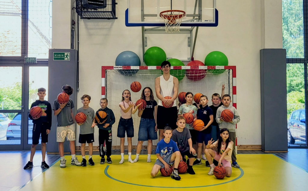Grupa uczniów stoi na szkolnej sali gimnastycznej z piłkami do koszykówki. W centrum znajduje się bardzo wysoki młody mężczyzna w białej koszulce sportowej, trzymający piłkę. Dzieci pozują wokół niego, część trzyma piłki, a jedna z uczennic podrzuca piłkę w górę. W tle widoczne są bramka, kosz do koszykówki oraz kolorowe piłki gimnastyczne. Zdjęcie wykonano podczas spotkania uczniów z absolwentem szkoły i koszykarzem.